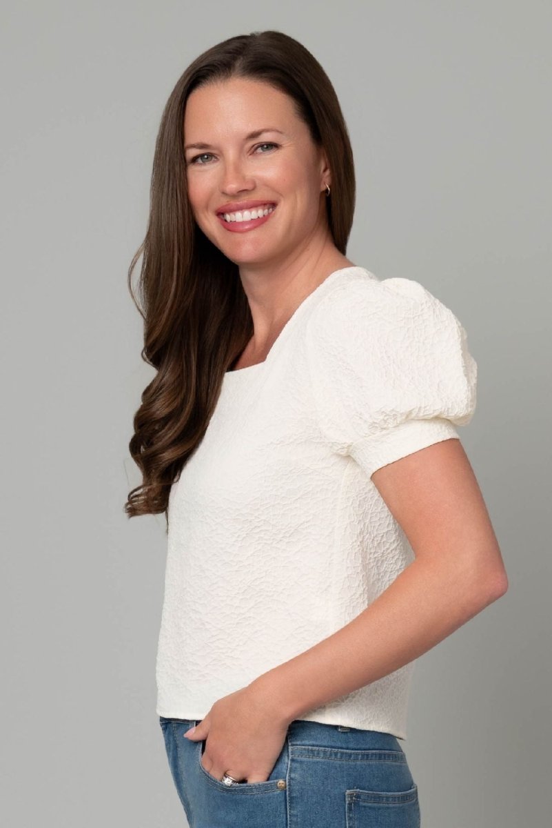 Woman wearing a white textured top and blue jeans against a gray background