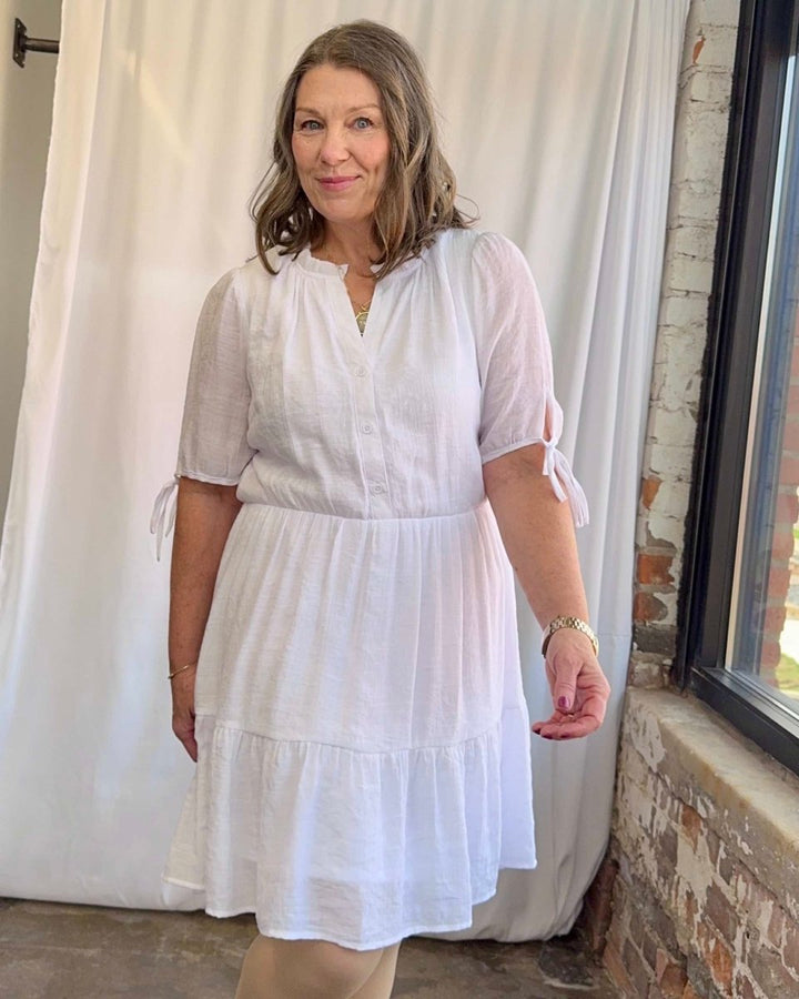 Woman wearing a white dress standing in front of a white curtain and brick wall.