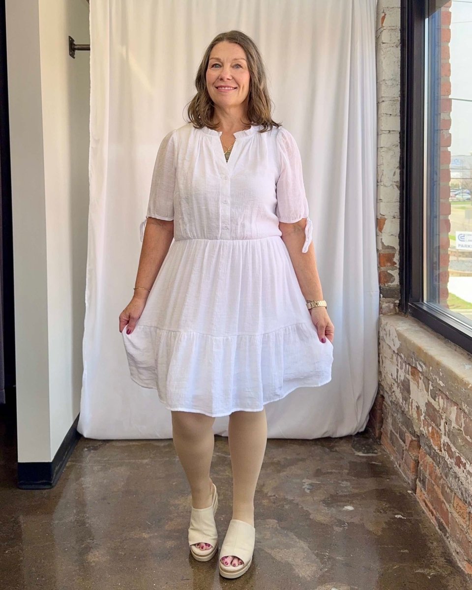 Woman wearing a white dress standing in a room with a window and brick wall.