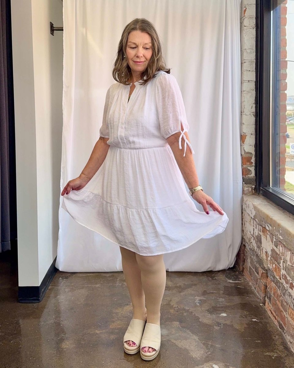 Woman wearing a white dress standing in a room with a white curtain and brick wall.