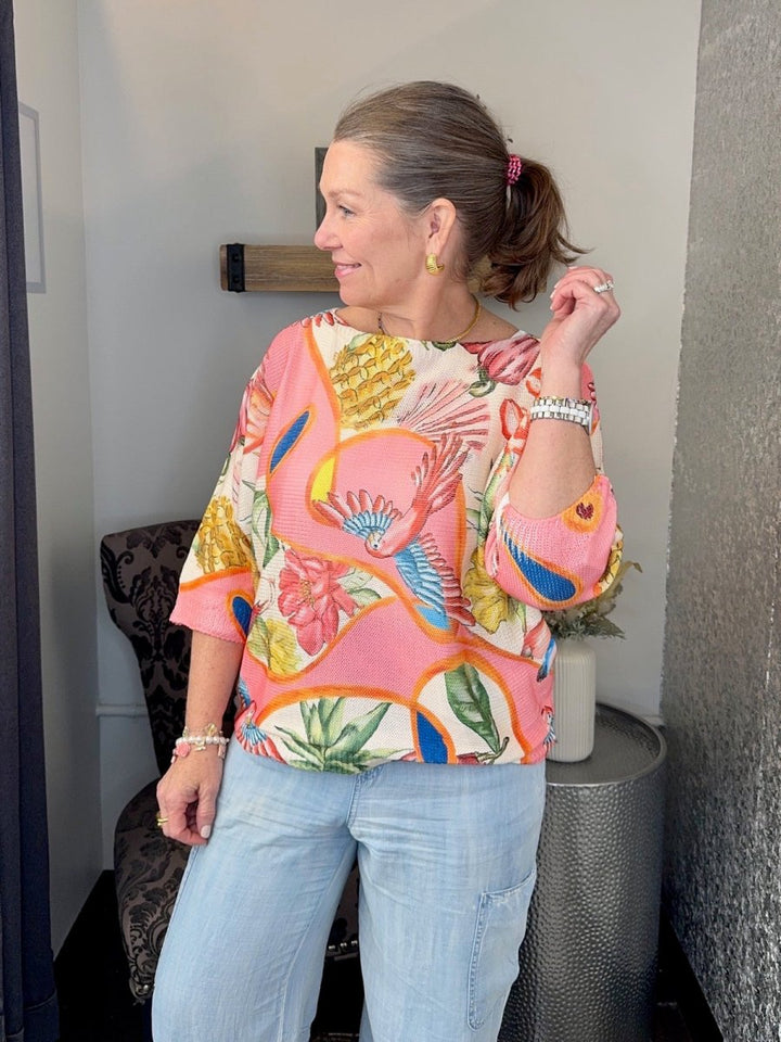 Woman wearing a Coral Multi tropical print batwing sweater with 3/4 sleeves and light wash denim jeans looking in a mirror.